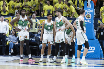 Four basketball players in white and green uniforms stand near the court as a yellow-clad crowd cheers in the background, mid-game scene.