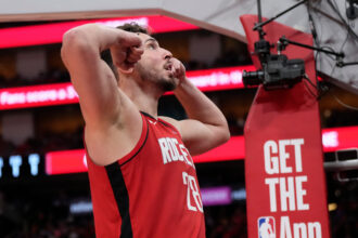 Male basketball player in a red jersey arms raised in triumph on the court, bright red signage in the background.