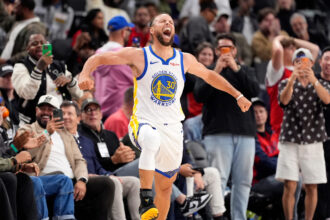 Stephen Curry of the Golden State Warriors celebrates on the court, arms outstretched, after a likely play, with fans cheering in the stands behind him.