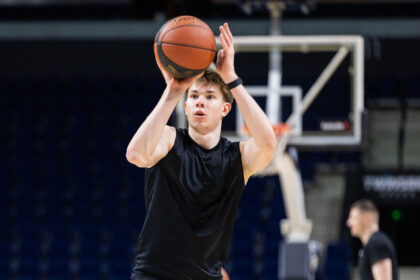 Basketball player in a black sleeveless shirt taking a jump shot indoors, ball raised above his head against a dark arena background