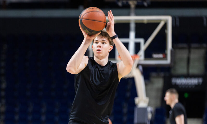 Basketball player in a black sleeveless shirt taking a jump shot indoors, ball raised above his head against a dark arena background