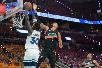 Basketball players leap at the rim; a white-jersey player (#30, RANDLE) blocks a shot by a Spurs opponent in black jersey near the hoop.