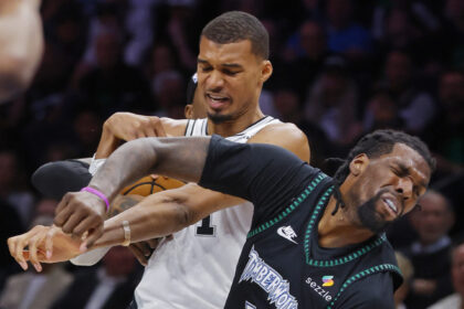 Two basketball players grapple for the ball during a game; one in white, one in black with teal trim, intense facial expressions nearby crowd Remain concise.