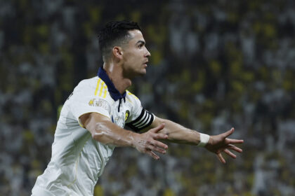 Soccer player in a white kit celebrating with arms outstretched, blurred stadium crowd in the background.