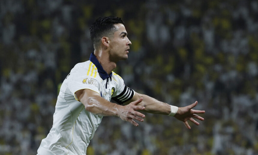 Soccer player in a white kit celebrating with arms outstretched, blurred stadium crowd in the background.
