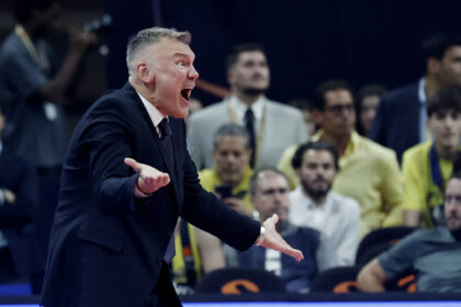 Man in a dark suit shouting with outstretched arms at a crowded sports event, audience in the background