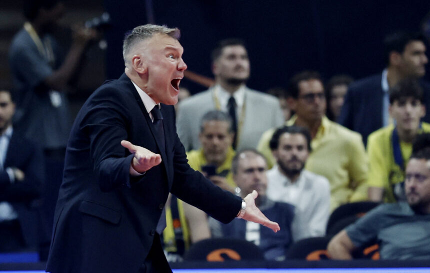 Man in a dark suit shouting with outstretched arms at a crowded sports event, audience in the background