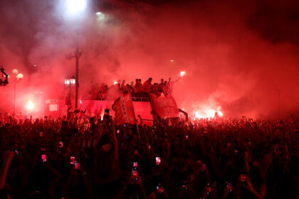 Crowd of fans at a nighttime victory celebration, red smoke fills the air while people on a raised float wave banners.