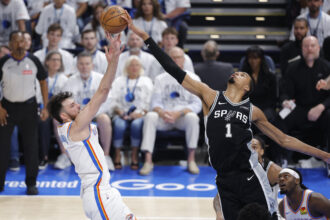 San Antonio Spurs guard in black jersey (number 1) reaches for a ball tipped by Oklahoma City Thunder player in a white jersey as they jump in an arena full of spectators.
