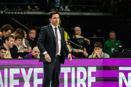 Basketball coach in a dark suit and tie standing courtside during a game, with players and spectators in the background.