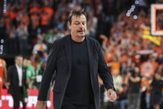 Man in a dark blazer walking on a basketball court, with a blurred crowd in orange cheering in the background.