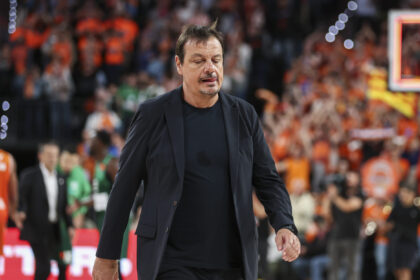Man in a dark blazer walking on a basketball court, with a blurred crowd in orange cheering in the background.