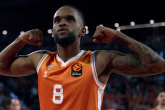 Basketball player in an orange Valencia jersey flexing his arms in celebration on the court, fists raised near his shoulders.