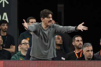 Man on stage with outstretched arms, shouting or speaking passionately, while a seated audience watches behind him.