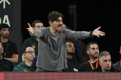 Man on stage with outstretched arms, shouting or speaking passionately, while a seated audience watches behind him.