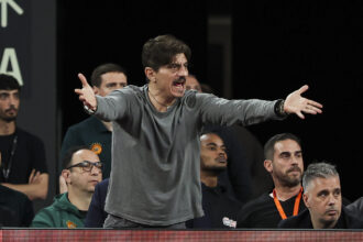 Man on stage with outstretched arms, shouting or speaking passionately, while a seated audience watches behind him.