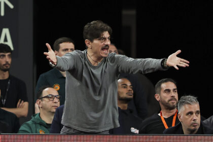 Man on stage with outstretched arms, shouting or speaking passionately, while a seated audience watches behind him.
