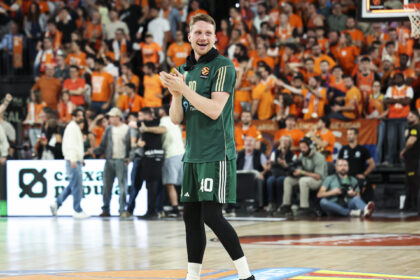Basketball player in a green jersey number 10 claps on the court as a cheering crowd in orange fills the stands behind him.
