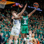 Two basketball players jump for the ball near the hoop as a defender closes in, with a crowded green-clad arena in the background.