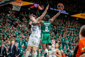 Two basketball players jump for the ball near the hoop as a defender closes in, with a crowded green-clad arena in the background.