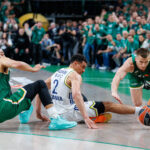 Basketball players in green and white uniforms dive for a loose ball as the crowd watches from the stands.