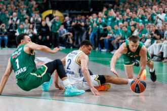 Basketball players in green and white uniforms dive for a loose ball as the crowd watches from the stands.