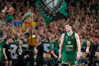 Basketball player in a green Zalgiris jersey #10 on the court as a packed arena cheers in the background, flag waving above the crowd.