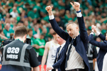 Man in a navy suit raises both arms in celebration as a green-clad crowd cheers in the background.