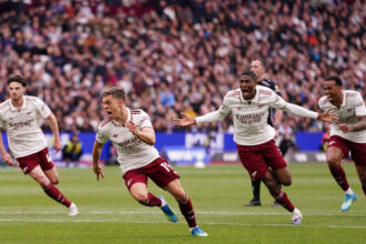 Arsenal players in white jerseys celebrate a goal on the pitch with a cheering crowd in the stands behind them.