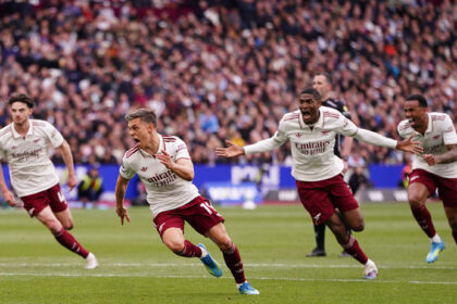 Arsenal players in white jerseys celebrate a goal on the pitch with a cheering crowd in the stands behind them.