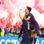 Two soccer players in navy uniforms celebrate a goal while pink confetti fills the stadium, fans cheering behind them.