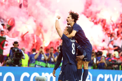 Two soccer players in navy uniforms celebrate a goal while pink confetti fills the stadium, fans cheering behind them.
