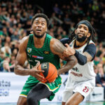 Two basketball players race for the ball during a match; a blurred crowd watches from the stands behind them.