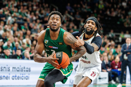 Two basketball players race for the ball during a match; a blurred crowd watches from the stands behind them.