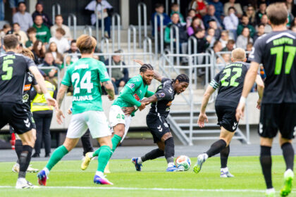 Soccer players in green and black jerseys chase the ball on a field with a crowded stands backdrop behind them.