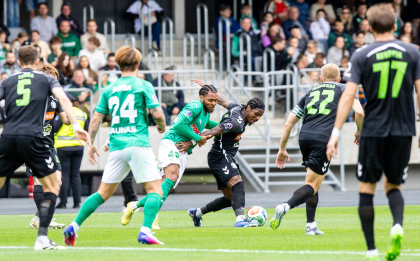 Soccer players in green and black jerseys chase the ball on a field with a crowded stands backdrop behind them.