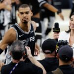 Basketball player in a Spurs jersey being interviewed by reporters at a press event.