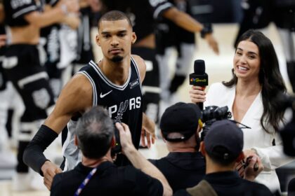 Basketball player in a Spurs jersey being interviewed by reporters at a press event.