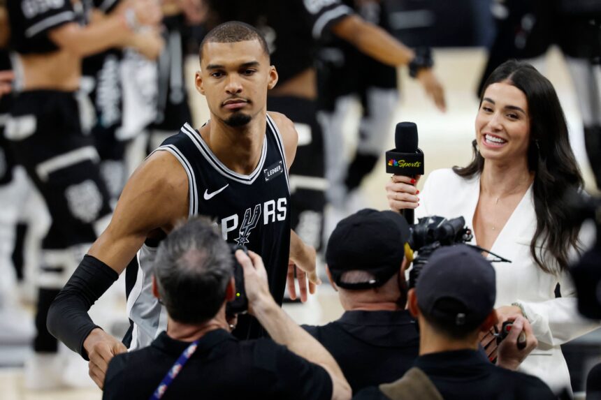Basketball player in a Spurs jersey being interviewed by reporters at a press event.