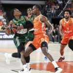 Basketball game: green-jersey player drives with the ball while an orange-jersey defender blocks him, crowd in background.