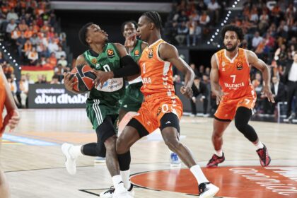 Basketball game: green-jersey player drives with the ball while an orange-jersey defender blocks him, crowd in background.