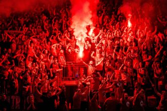 Dense crowd of sports fans cheering in a stadium, with red flare light and smoke in the center.