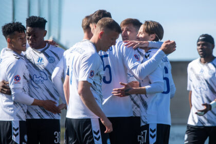 Soccer players in white jerseys form a celebratory huddle on a sunny training field.