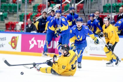 Ice hockey action: a yellow-team player falls on the ice while pursuing the puck, with blue-team players nearby and spectators in the stands behind.