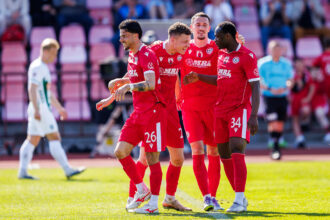 Four soccer players in red uniforms celebrate together on the sunny grass of a stadium field, arms around each other in a cheer.