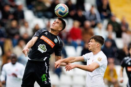 Two soccer players leap for a header as a colorful ball hovers above them on the field with a blurred crowd in the background, 0-0 score banner below