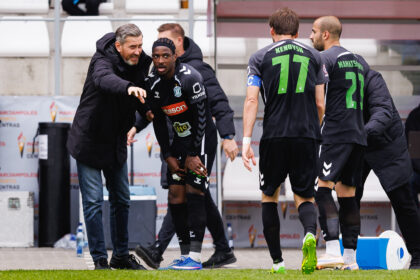 Soccer players in black kits stand near the sideline as a coach points and talks to them.