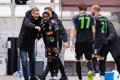 Soccer players in black kits stand near the sideline as a coach points and talks to them.