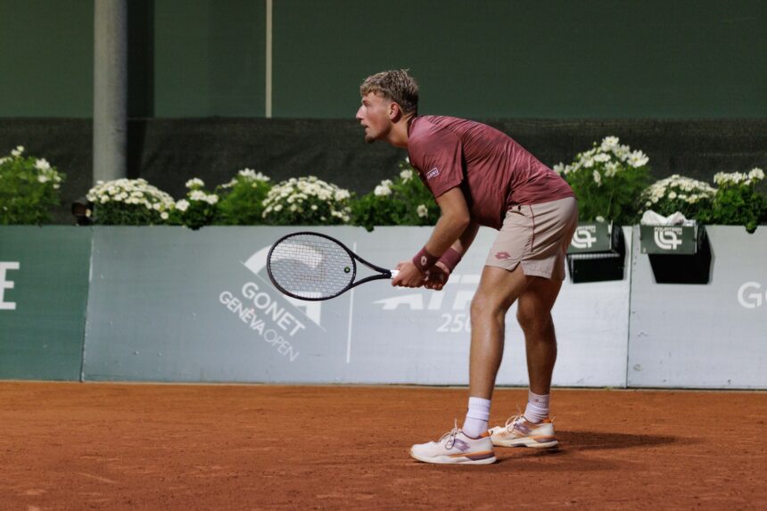 Tennis player in a maroon shirt bending forward, preparing to hit a shot on a clay court.