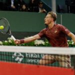 Male tennis player in a maroon shirt lunges to hit a yellow ball with a racket on an indoor court, spectators in the stands behind him.